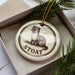 A ceramic ornament in a box, featuring a small illustration of a ferret.