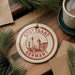A circular ornament with the text "STUTTGART GERMANY" is displayed on a wooden surface, surrounded by pine branches and a cup of coffee.