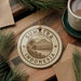 a decorative ornament with the text "SUMATRA INDONESIA" on it, surrounded by pine branches and a cup of coffee on a wooden surface.