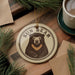 A circular ornament featuring a bear image is displayed on a wooden surface, surrounded by pine branches and a cup of coffee.
