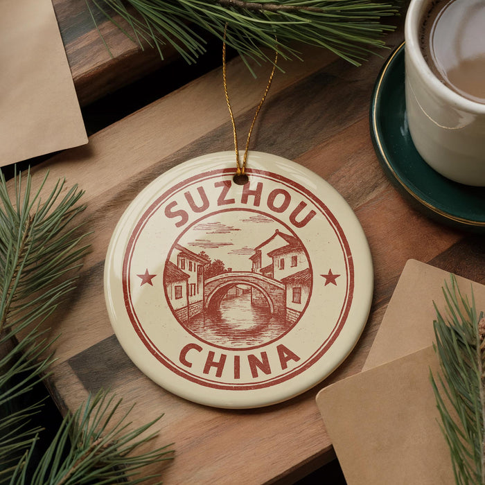 A round ornament with the text "SUZHOU CHINA" and a drawing of a bridge is displayed on a wooden surface, surrounded by pine branches and a cup of coffee.