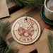 A round ornament with the text "SUZHOU CHINA" and a drawing of a bridge is displayed on a wooden surface, surrounded by pine branches and a cup of coffee.
