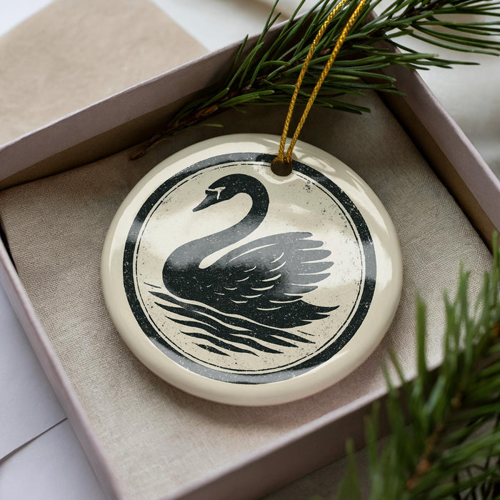 A ceramic ornament in the shape of a swan, with a black and white design, is displayed in a box with pine branches.