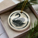 A ceramic ornament in the shape of a swan, with a black and white design, is displayed in a box with pine branches.