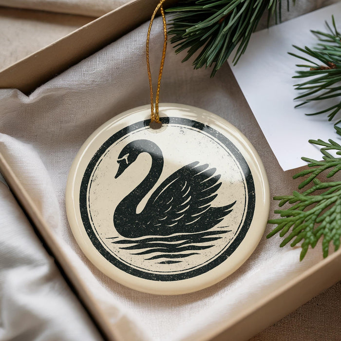 A decorative ornament in the shape of a swan, featuring a black silhouette against a white background, is displayed in a box with pine branches.