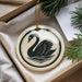 A decorative ornament in the shape of a swan, featuring a black silhouette against a white background, is displayed in a box with pine branches.