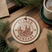 A circular ornament with the text "SZEGED HUNGARY" and an illustration of a city skyline hangs on a wooden surface, surrounded by pine branches and a cup of coffee.