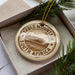 A decorative ornament in a box, featuring a circular design with text and an illustration of a mountain landscape.
