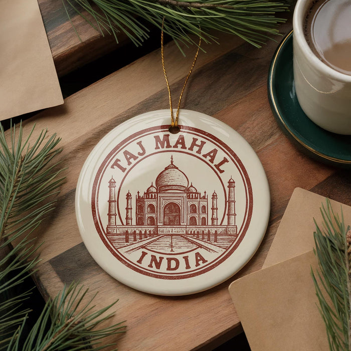 A circular ornament with the text "Taj Mahal, INDIA" printed on it, featuring an illustration of the Taj Mahal. The ornament is hanging from a string, and there is a cup of coffee and some pine branches in the background.