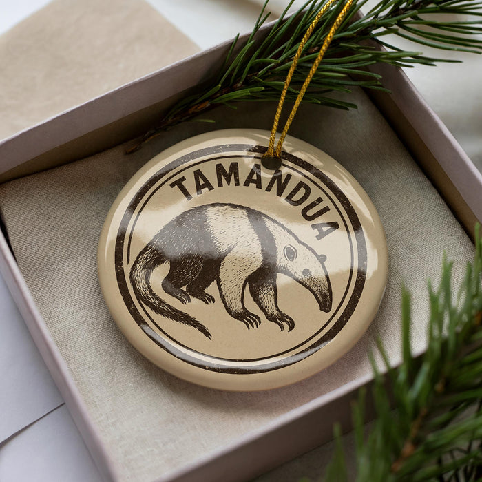 A circular ornament with a printed image of a tamandua, a type of primate, and the word "TAMANDU" written on it, is displayed in a box with pine branches.