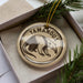 A circular ornament with a printed image of a tamandua, a type of primate, and the word "TAMANDU" written on it, is displayed in a box with pine branches.