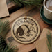 A circular ornament featuring a graphic of a Tamandua, a type of rodent, is displayed on a wooden surface alongside a cup of coffee and some pine branches.