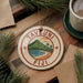 A round ornament with the text "TAVEUNI Fiji" and a tropical landscape design, placed on a wooden surface next to a cup of coffee and pine branches.