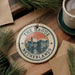 A decorative ornament with the text "THE RAGUE NETHERLANDS" and an illustration of a cityscape, placed on a wooden surface alongside a cup of coffee and pine branches.