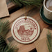 A circular ornament with the text "THIRUVANANTHAPURAM INDIA" printed on it, surrounded by pine branches and a cup of coffee on a wooden surface.