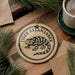 A round ornament featuring a tiger salamander design, placed on a wooden surface with pine branches and a cup of coffee nearby.