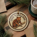 A circular ornament featuring a kangaroo is displayed on a wooden surface, surrounded by pine branches and a cup of coffee.