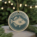 A circular ornament with the word "TRIGGERFISH" printed on it, featuring a fish illustration. The ornament is hanging from a string, and there are pine branches and lights in the background.