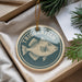 A round ornament with a fish design, hanging from a string, placed inside a box with pine branches.