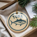 A round ornament with a fish design and the word "Tuna" printed on it, hanging from a string.