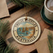 A decorative ornament featuring a scenic image of the Twelve Apostles, a famous limestone formation in Australia, is displayed on a wooden surface alongside a cup of coffee and some pine branches.