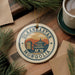 A circular ornament with the text "ULANBAATAR MONGOLIA" on it, surrounded by pine branches and a cup of coffee on a wooden surface.