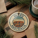 A circular ornament with the text "ULORU AUSTRALIA" and an illustration of Uluru, a famous rock formation in Australia, is displayed on a wooden surface alongside a cup of coffee and pine branches.
