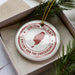 A white ornament with a red border and a map of St. Vincent and the Grenadines, placed in a box with pine branches.