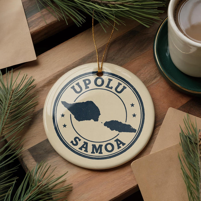 A circular ornament with the text "UPOLU SAMOA" and a map of the island of Samoa, placed on a wooden surface next to a cup of coffee and pine branches.
