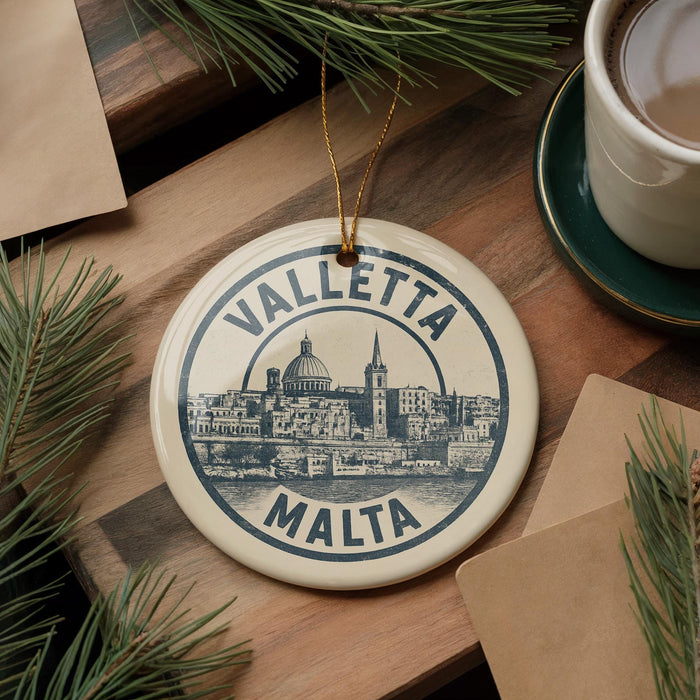 A circular ornament with the text "VALLETA MALTA" and an illustration of a cityscape is displayed on a wooden surface, surrounded by pine branches and a cup of coffee.
