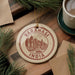 A circular ornament with the text "VARANASI INDIA" printed on it, surrounded by pine branches and a cup of coffee on a wooden surface.