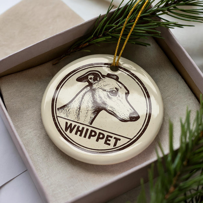 A ceramic ornament in the shape of a dog's head, with the word "WHIPPET" written below it, is displayed in a box with pine branches.