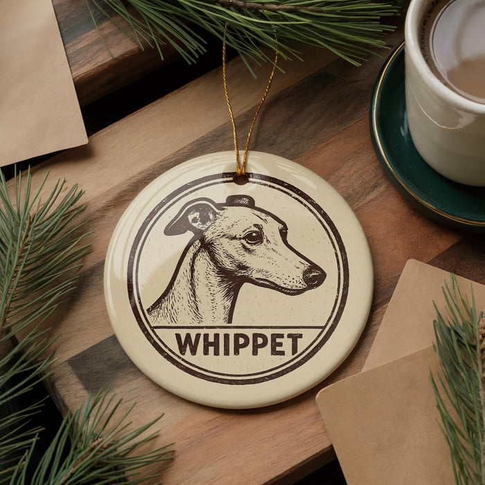 A round ornament featuring a Whippet dog's head, with the word "Whippet" written below it, is displayed on a wooden surface alongside a cup of coffee and some pine branches.