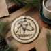 A round ornament featuring a Whippet dog's head, with the word "Whippet" written below it, is displayed on a wooden surface alongside a cup of coffee and some pine branches.