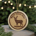 A round ornament with a deer image and the text "WHITE-TAILED DEER" on it, hanging in front of a Christmas tree with lights.