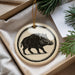 A decorative ornament featuring a black and white illustration of a wild boar is displayed in a box, surrounded by pine branches.