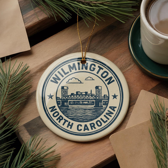 A circular ornament with the text "WILMINGTON NORTH CAROLINA" and an illustration of a bridge, placed on a wooden surface next to a cup of coffee and some pine branches.