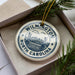 A decorative ornament in a box, featuring a circular design with a city skyline and water, surrounded by greenery.