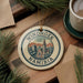 A decorative ornament with the text "WINDHOOK NAMIBIA" on it, featuring an illustration of Namibia's capital Windhoek. The ornament is placed on a wooden surface, surrounded by pine branches and a cup of coffee.