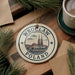 A circular ornament with the text "Wrocław Poland" and an illustration of a city skyline hangs on a wooden surface, surrounded by pine branches and a cup of coffee.