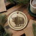 a ceramic ornament with a detailed illustration of a yak, a large, shaggy-haired animal, on a wooden surface. The ornament is hanging from a string, and there is a cup of coffee and some pine branches in the background.