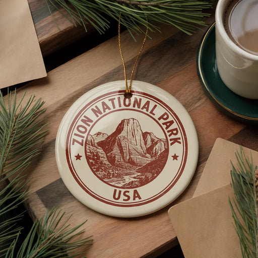 A circular ornament with the text "ZION NATIONAL PARK USA" printed on it, featuring a mountain landscape design, is displayed on a wooden surface alongside a cup of coffee and some pine branches.