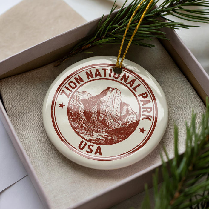 A white ornament with a red border, featuring an illustration of a mountain range and the text "ZION NATIONAL PARK USA" on it.