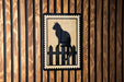A black silhouette of a cat sitting on a wooden fence is displayed on a wooden wall.