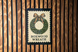 A sign with a wreath design and the text "BOXWOOD WRATH" is displayed on a wooden wall.