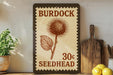 A vintage-style seedhead sign with the text "Burdock 30c Seedhead" is displayed on a shelf next to a potted plant and a wooden cutting board.