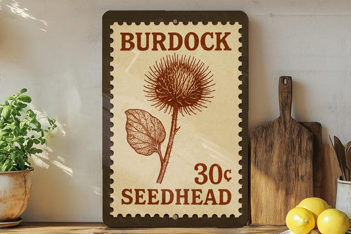 A vintage-style seedhead sign with the text "Burdock 30c Seedhead" is displayed on a shelf next to a potted plant and a wooden cutting board.