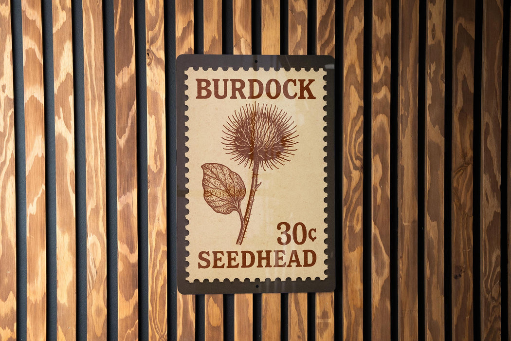 A wooden wall with a postage stamp-like sign that reads "Burdock 30c Seedhead" attached to it.