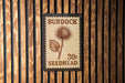 A wooden wall with a postage stamp-like sign that reads "Burdock 30c Seedhead" attached to it.