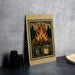 A metal sign with a wooded scene and a campfire, featuring two mugs, is displayed on a countertop.